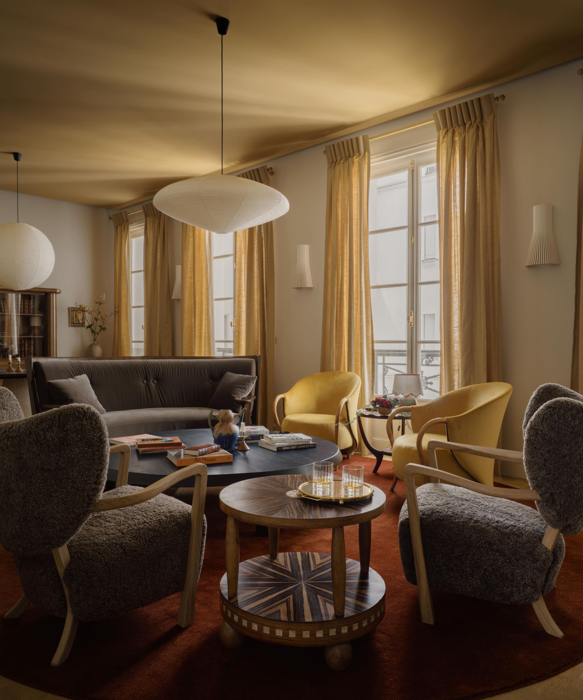 A living room with an ochre ceiling, warm white walls, and a large window with ochre curtains. A dark red rug and dark neutral chairs.