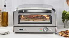 A Cuisinart Indoor Pizza Oven on a white kitchen countertop, cooking a pizza with a cooked pizza next to it