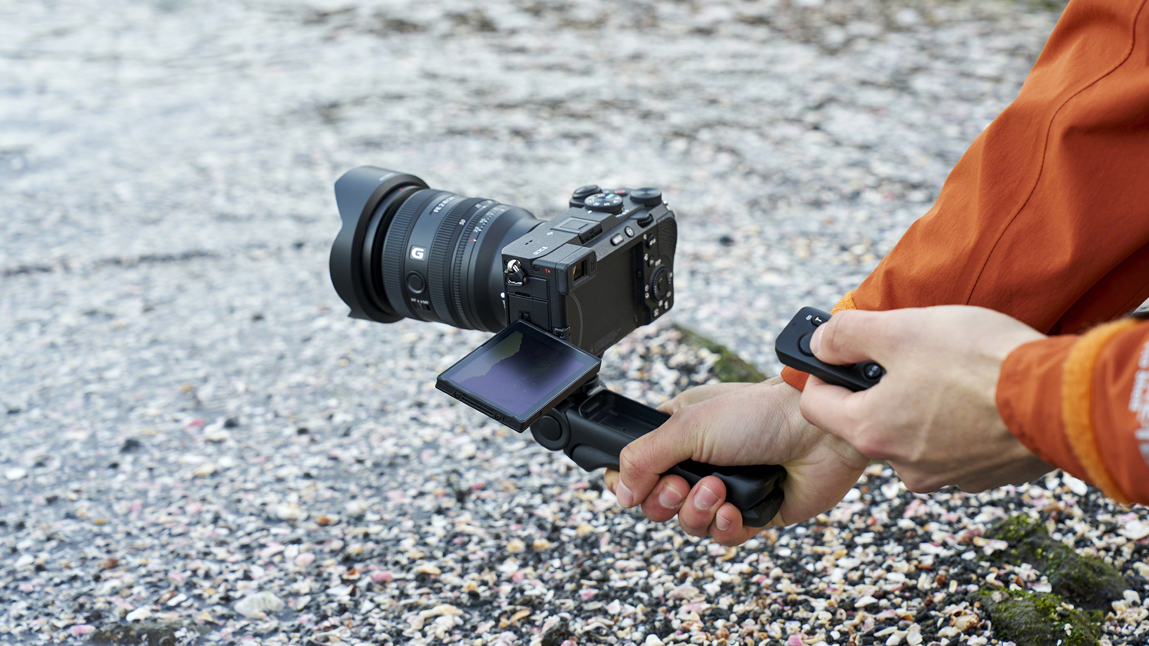 A person in an orange jacket holds a camera with with GP-VPT3 grip, a flip screen and Sony RMT-VP2 remote control, standing on a pebbled beach by the water
