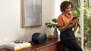 The Sonos Era 300 pictured on a wooden cabinet while a woman leans against it and uses her smartphone