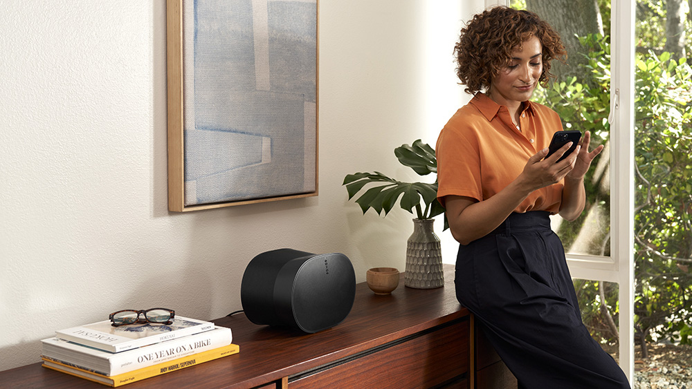 The Sonos Era 300 pictured on a wooden cabinet while a woman leans against it and uses her smartphone