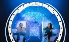 A futuristic-like vehicle with white seats and white hand rest. A man sitting on the chair on the left and a woman sitting on a chair on the right. Photographed against a blue background