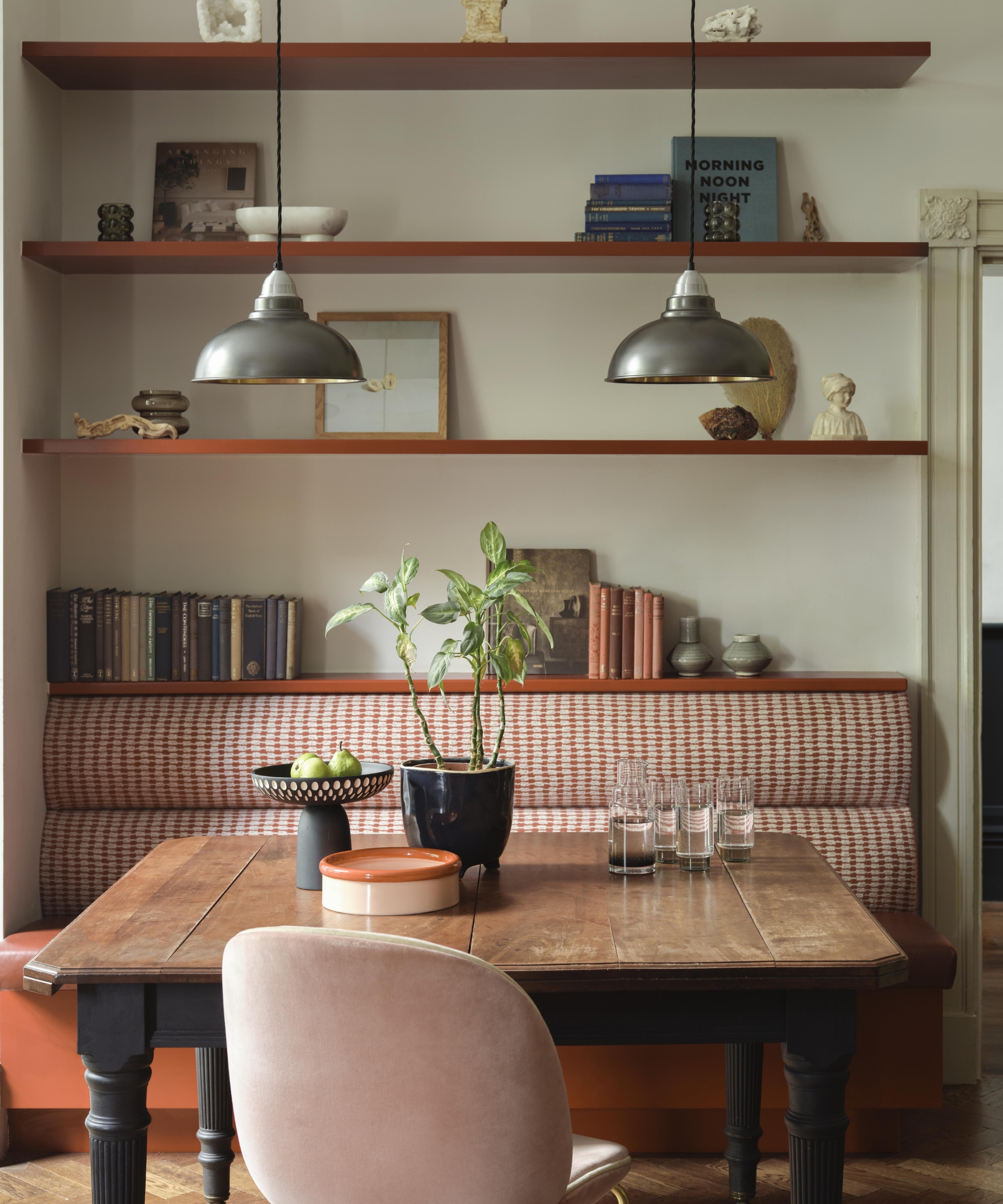 Dining nook with a banquette seat, wooden table, open shelving of books and ornaments, and pendant lamps.