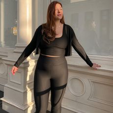 Mid-size fashion influencer Raeann Langas poses against a building in NYC wearing a workout outfit cropped zip-up hoodie, black sports bra, and black leggings