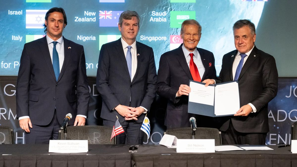 four men in dark suits stand in front of a table with microphones and small flags on it.