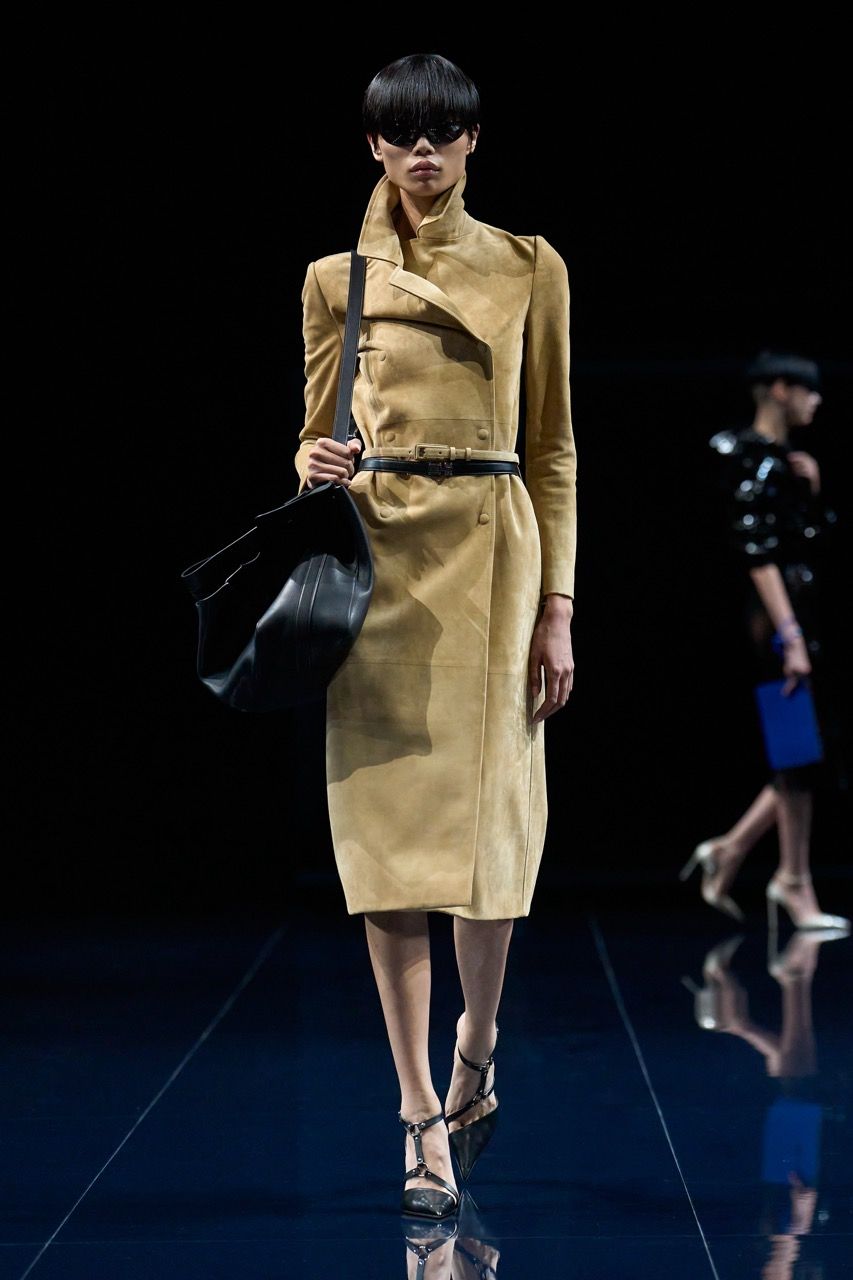 a model at Tom Ford Spring 2026 wearing a funnel-neck tan suede trench coat, two thin belts, a black bag, and strappy black heels