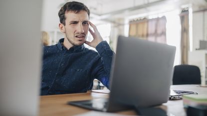 A young man looks perplexed as he looks at his laptop while sitting at his desk.