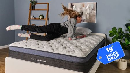 The image shows a light haired mattress tester diving onto the Helix Midnight Luxe mattress, placed on a white bed frame in a neutrally decorated bedroom. Inset, a badge reads 'tg price drop'