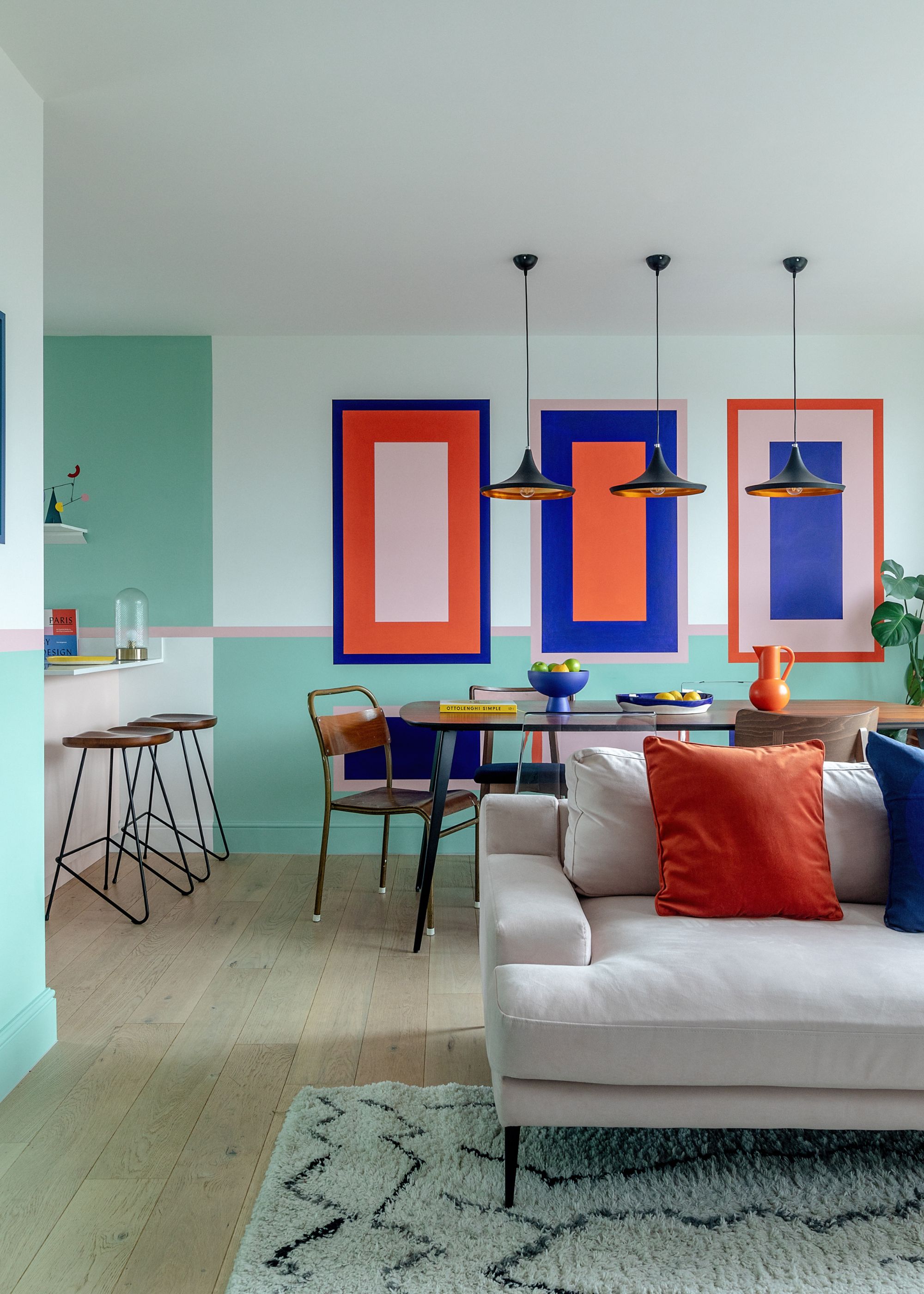 A modern living and dining space with geometric shapes painted onto the walls in a palette of mint green, bright red, bubblegum pink, and electric blue. Wood flooring with a taupe sofa and cream patterned rug.
