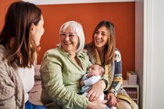 Happy grandmother with grey hair smiling while holding her grandson next to her daughters in a bedroom