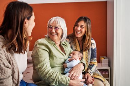 Happy grandmother with grey hair smiling while holding her grandson next to her daughters in a bedroom