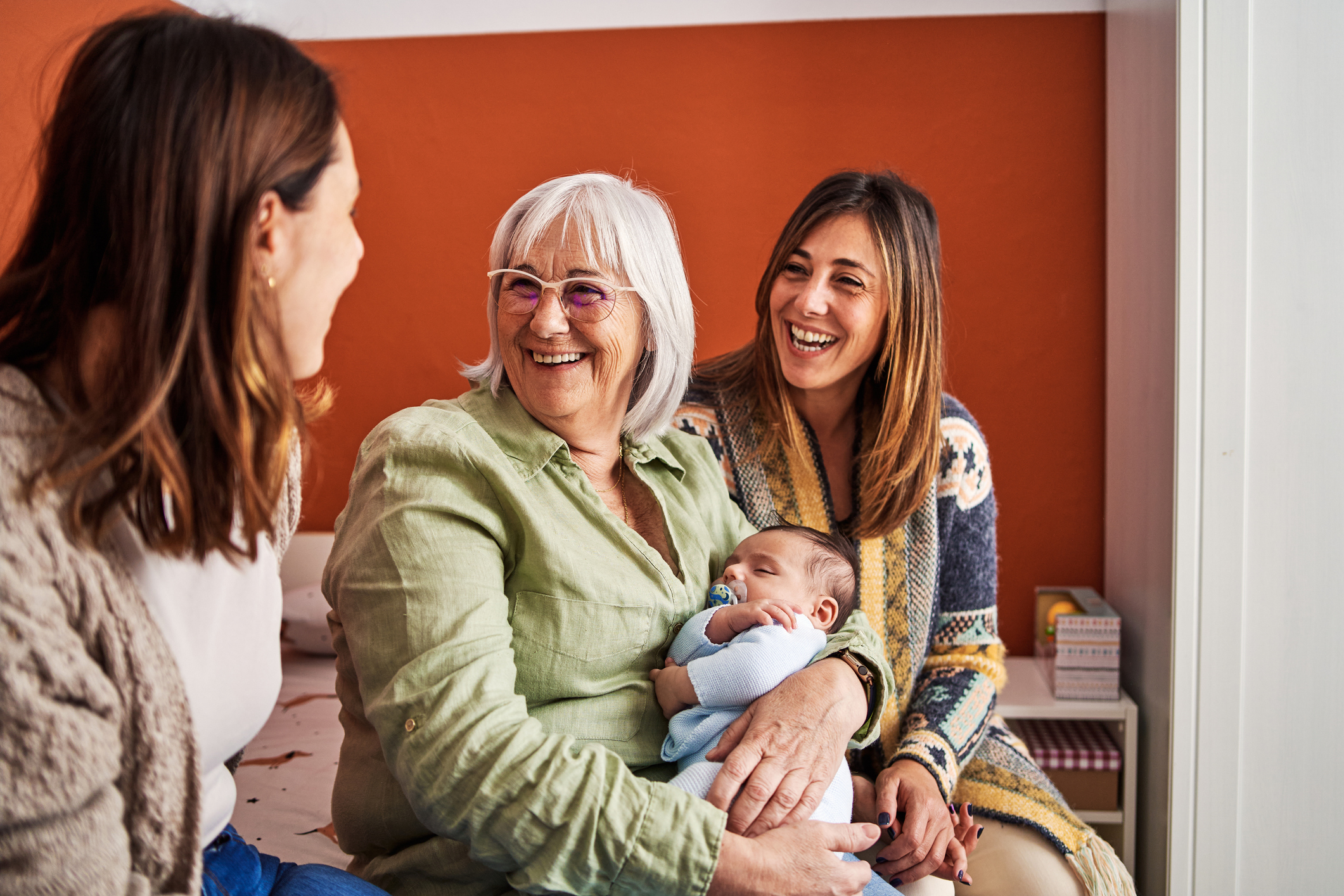 Happy grandmother with grey hair smiling while holding her grandson next to her daughters in a bedroom