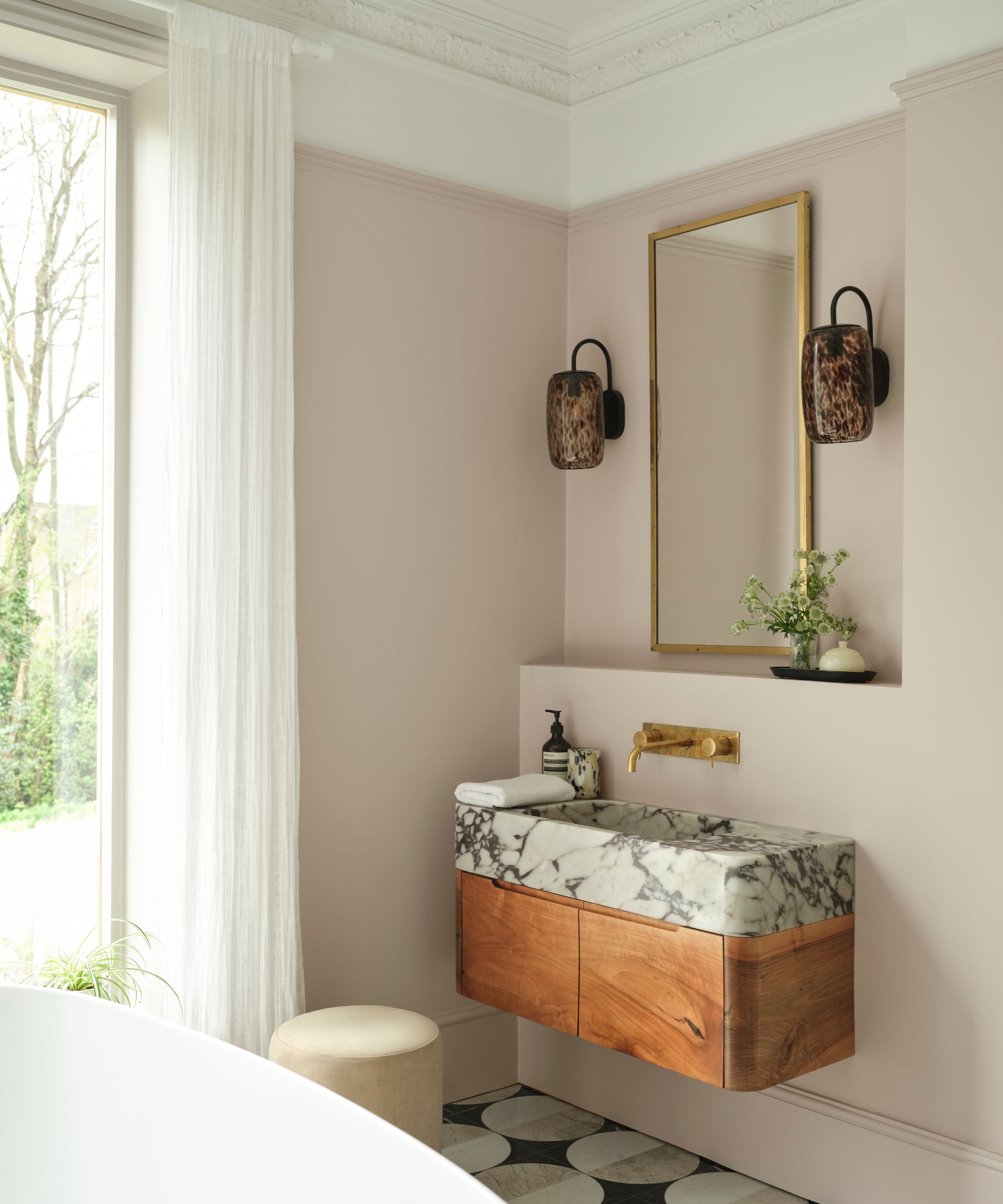 A modern bathroom with soft pink walls, a wooden vanity with a marble sink, a brass wall mirror, tiled flooring, and a floor-to-ceiling window with white sheer curtains.
