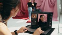 Photographer editing a portrait of a woman on a laptop in a studio with pink draped background and model in the background