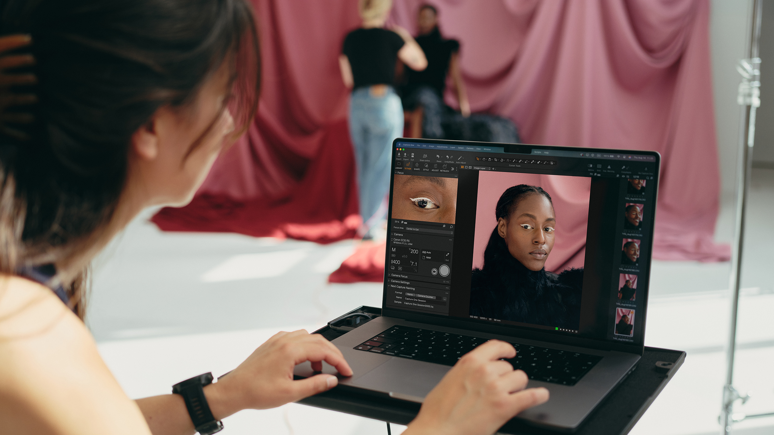 Photographer editing a portrait of a woman on a laptop in a studio with pink draped background and model in the background