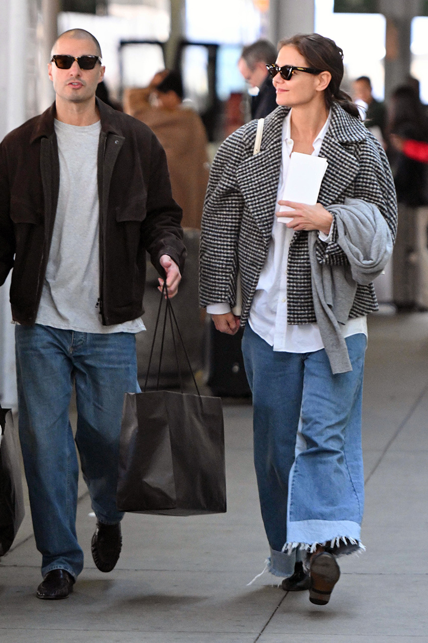 Katie Holmes at JFK airport wearing a printed black-and-white checked coat over a white button-down shirt with wide-leg jeans and black loafers