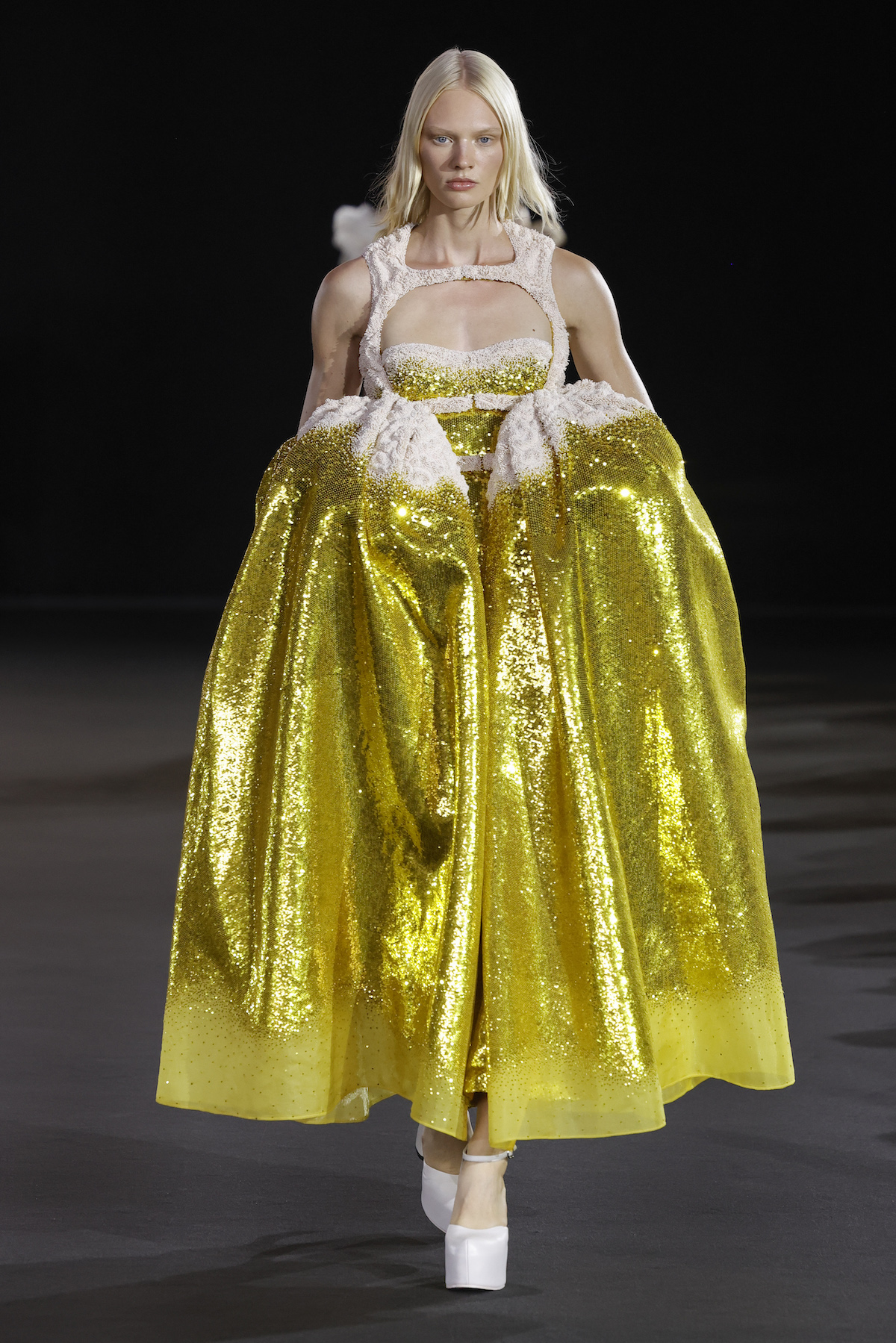 A female model wearing a long sleeveless white and gold dress.