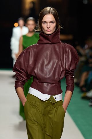 A photo from the Victoria Beckham S/S 25 runway show of a model wearing a burgundy leather high-neck top with a fitted bodice and oversized sleeves and olive pants.