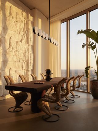 A dining room flooded with golden light through floor to ceiling windows, a wooden dining table with woven sea grass chairs