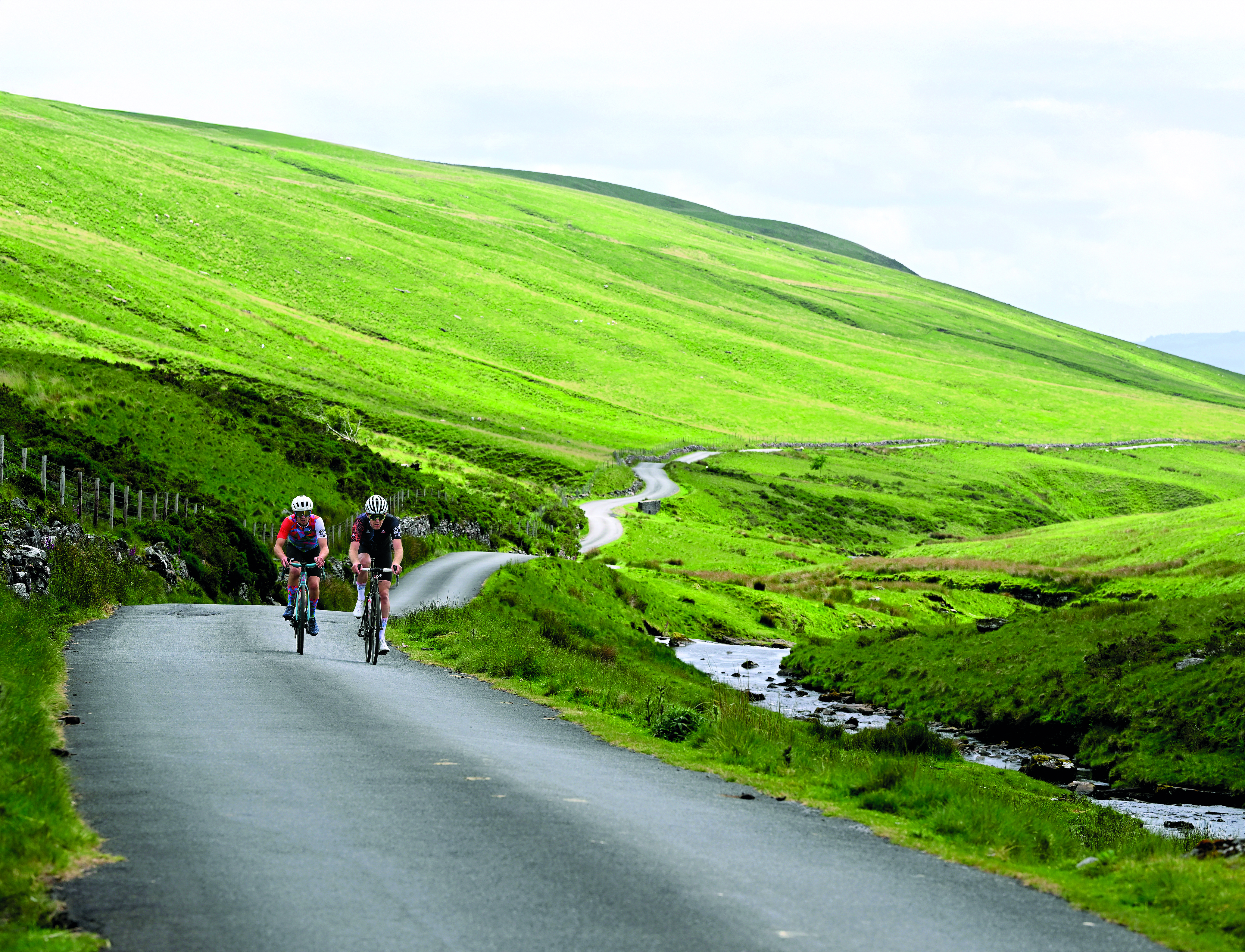 Two riders climb towards the camera with a ribbon like road behind them and fields either side