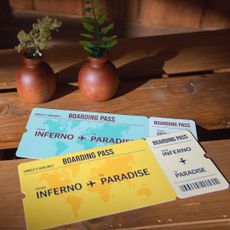 A close-up of a wooden table in a shed, with a wooden barrel in the corner. Yellow and blue boarding passes lay on the table, reading "Single's Airlines. Inferno to Paradise." A promo image from Netflix's 'Single's Inferno' season 5.