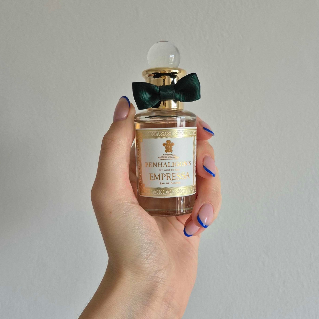 A close-up of Digital Beauty Writer Naomi Jamieson's hand, seen holding a bottle of Penhaligon's Empressa perfume, which features a dark green bow/ in front of a cream wall background