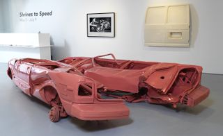 A squashed peach coloured car, vertically split in two, displayed on a grey floor of an art gallery. On the white walls are framed art and card parts