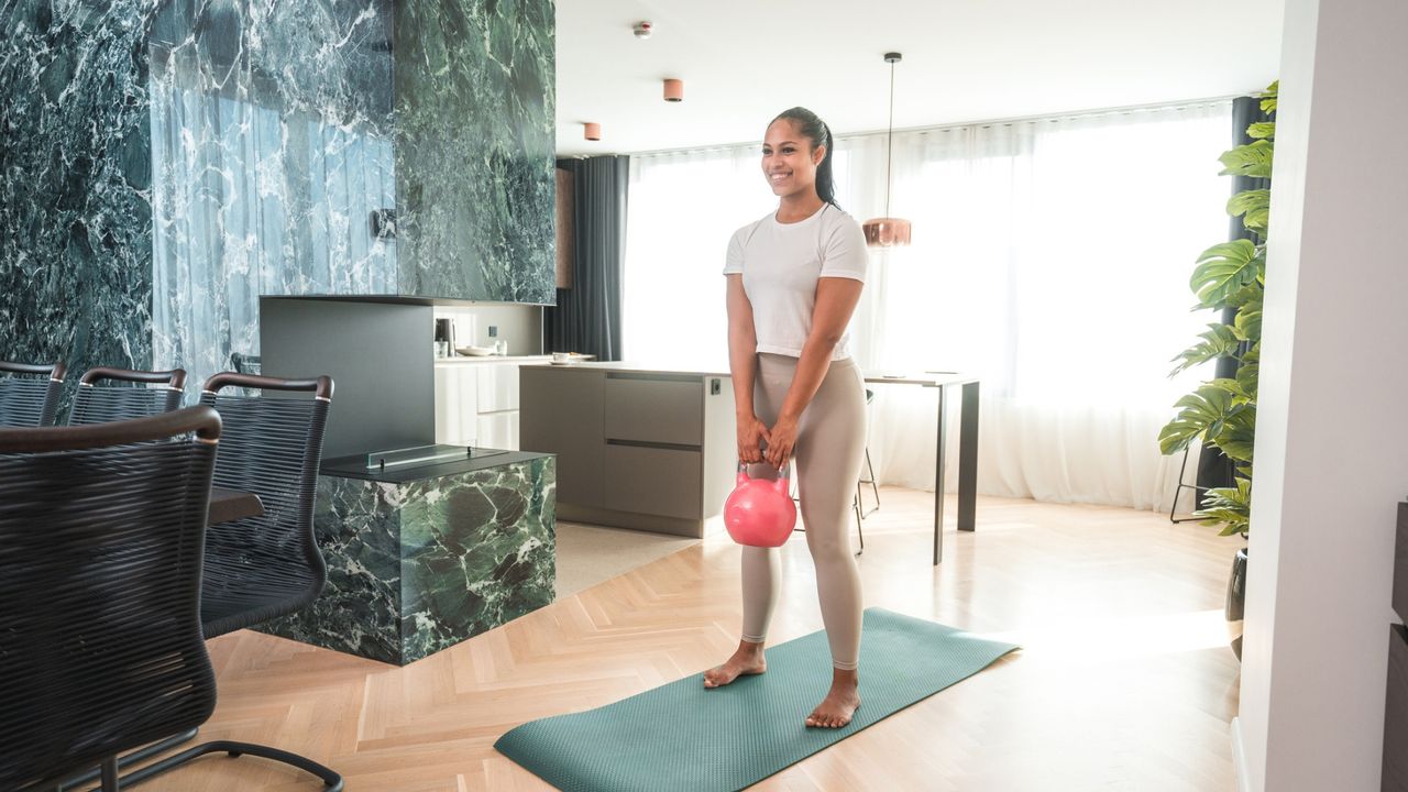 woman in leggings and tshirt standing on an exercise mat holding a pink kettlebell in front of her. she&#039;s in a white and green living room setting. 