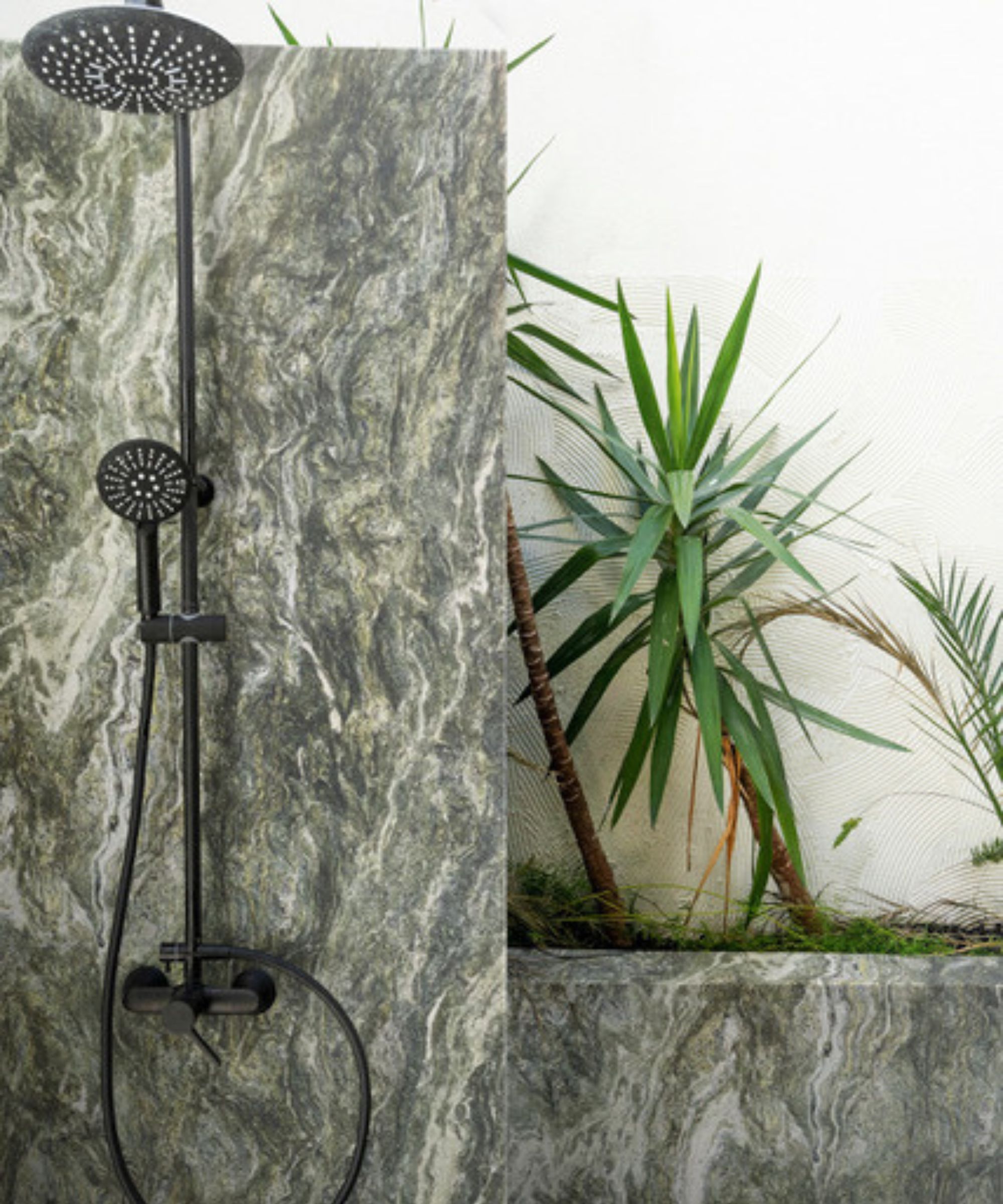 A black rainfall shower and handheld fitting mounted on a large format green marble panel, beside a yucca plant and rendered wall.