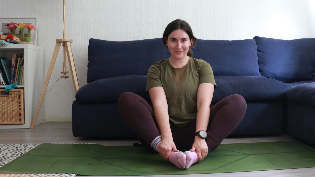 Marina Leiva Garc&iacute;a performs a butterfly pose at home. She is sitting with her knees bent and the soles of her feet together. Her knees are pointing upwards at a 45&deg; angle. Behind her is a couch.