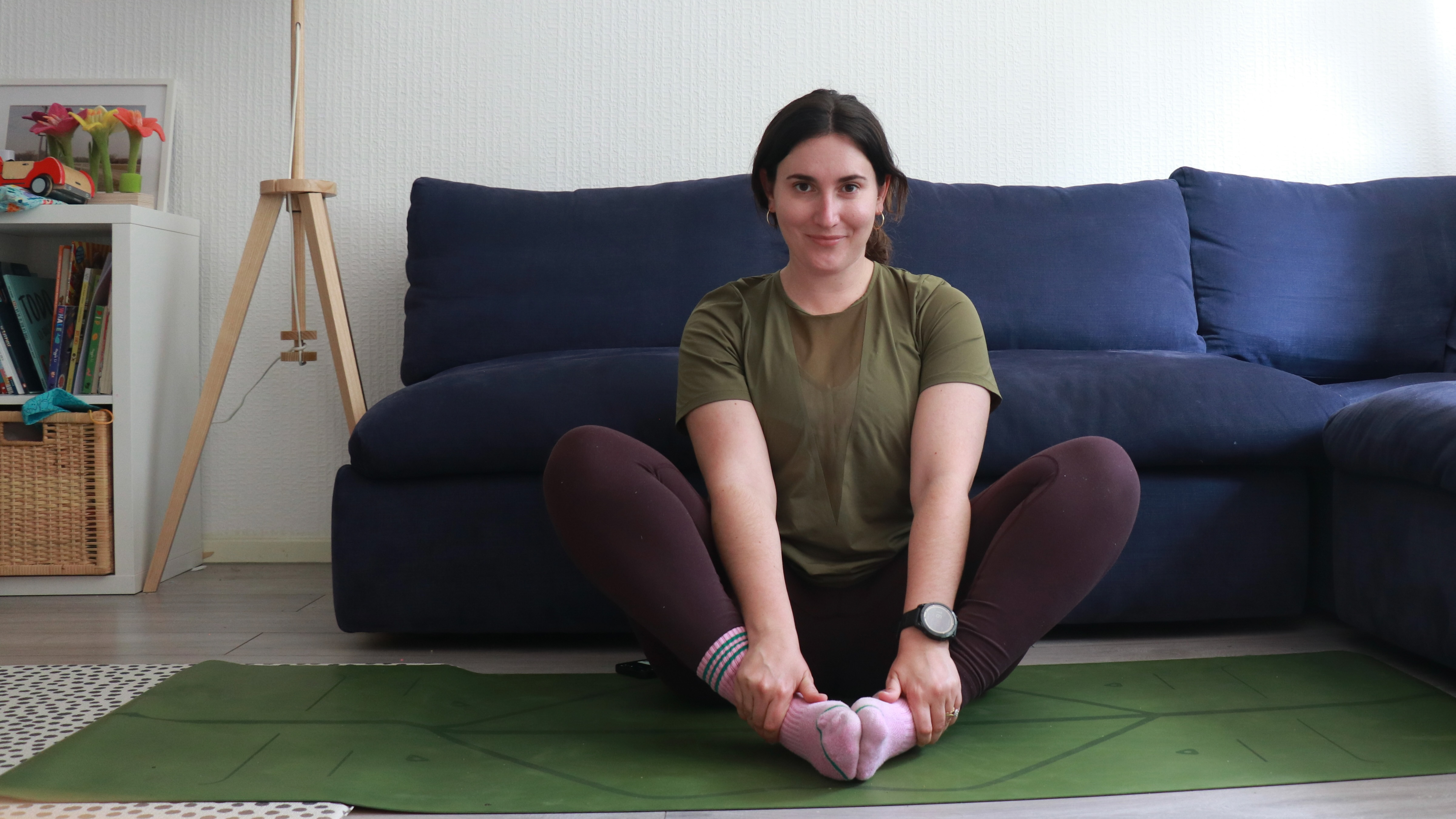 Marina Leiva García performs a butterfly pose at home. She is sitting with her knees bent and the soles of her feet together. Her knees are pointing upwards at a 45° angle. Behind her is a couch.