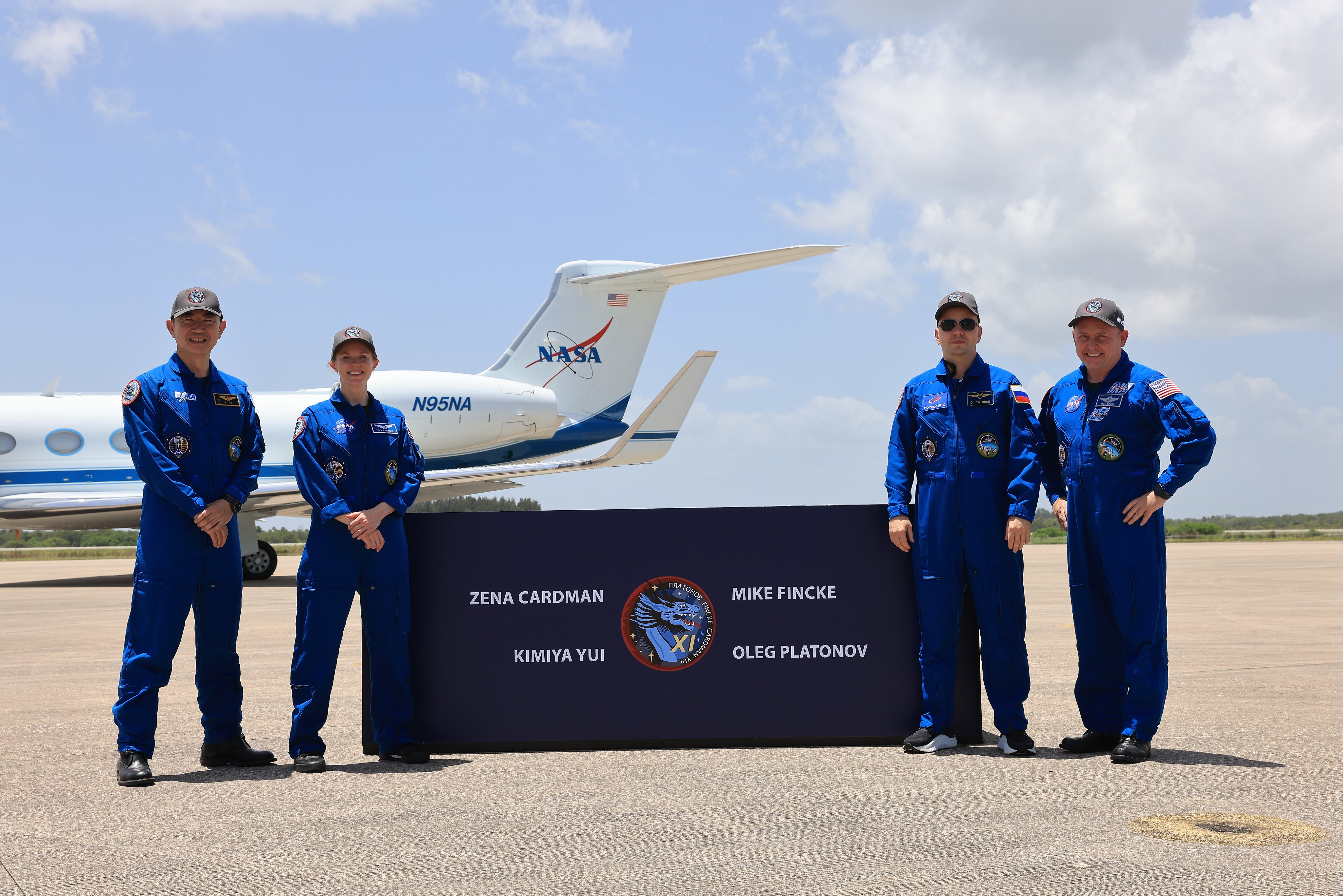 Four people in blue jumpsuits stand two on either side of a sign with a mission patch and their names on it.