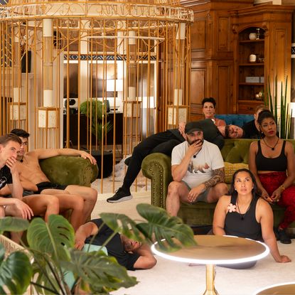 A group of people (L-R: ATHENA V, NICK, SHANE, STEVE, VAL, SUSAN, ATHENA S, OMAROSA) sit on green couches in a room decorated with plants, circular tables, and a life-sized gold cage, in Hulu's 'Got to Get Out'