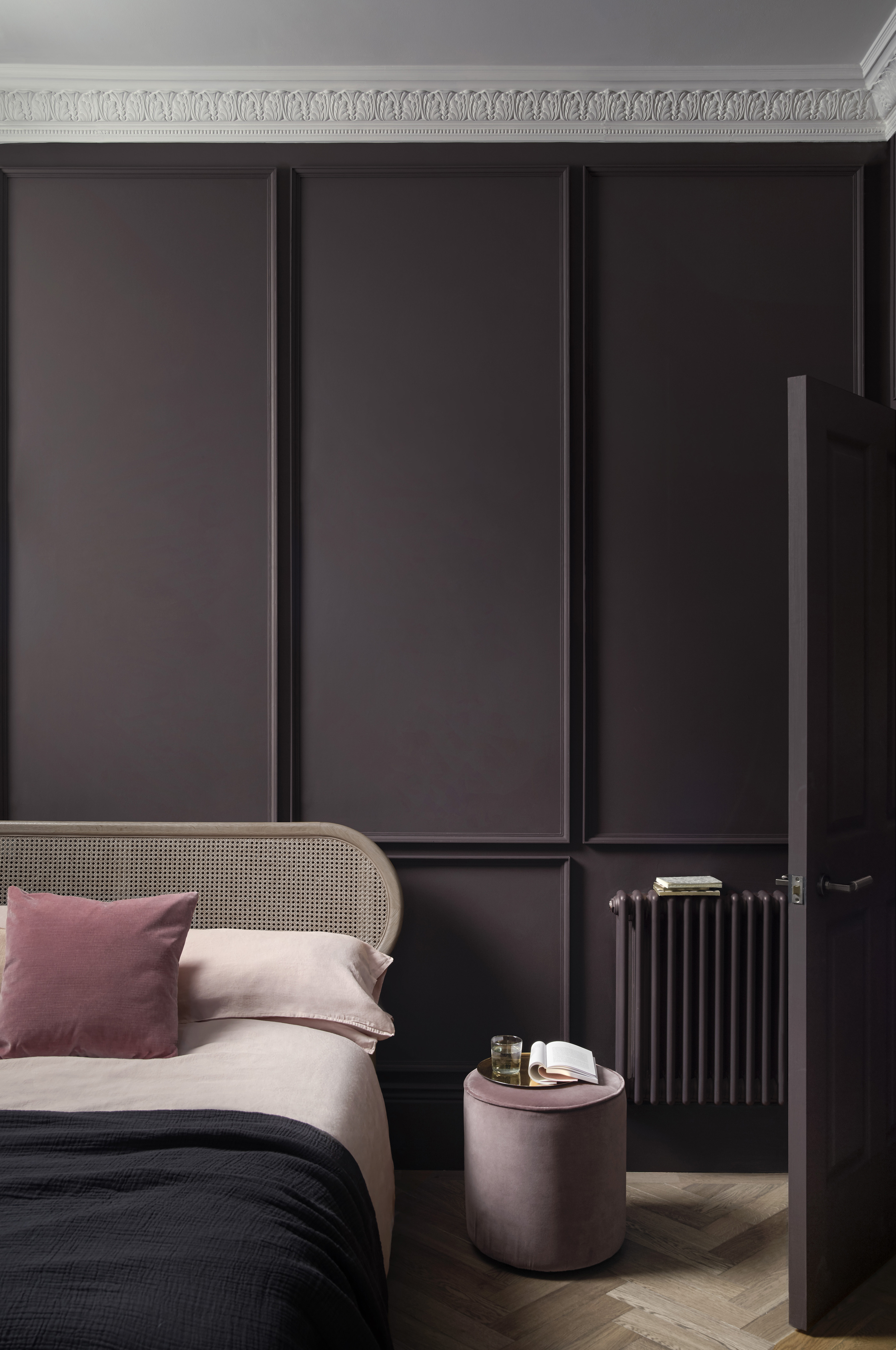 A deep aubergine scheme demonstrating the best bedroom colors, with panelled walls, a rattan bed and pale pink accessories.
