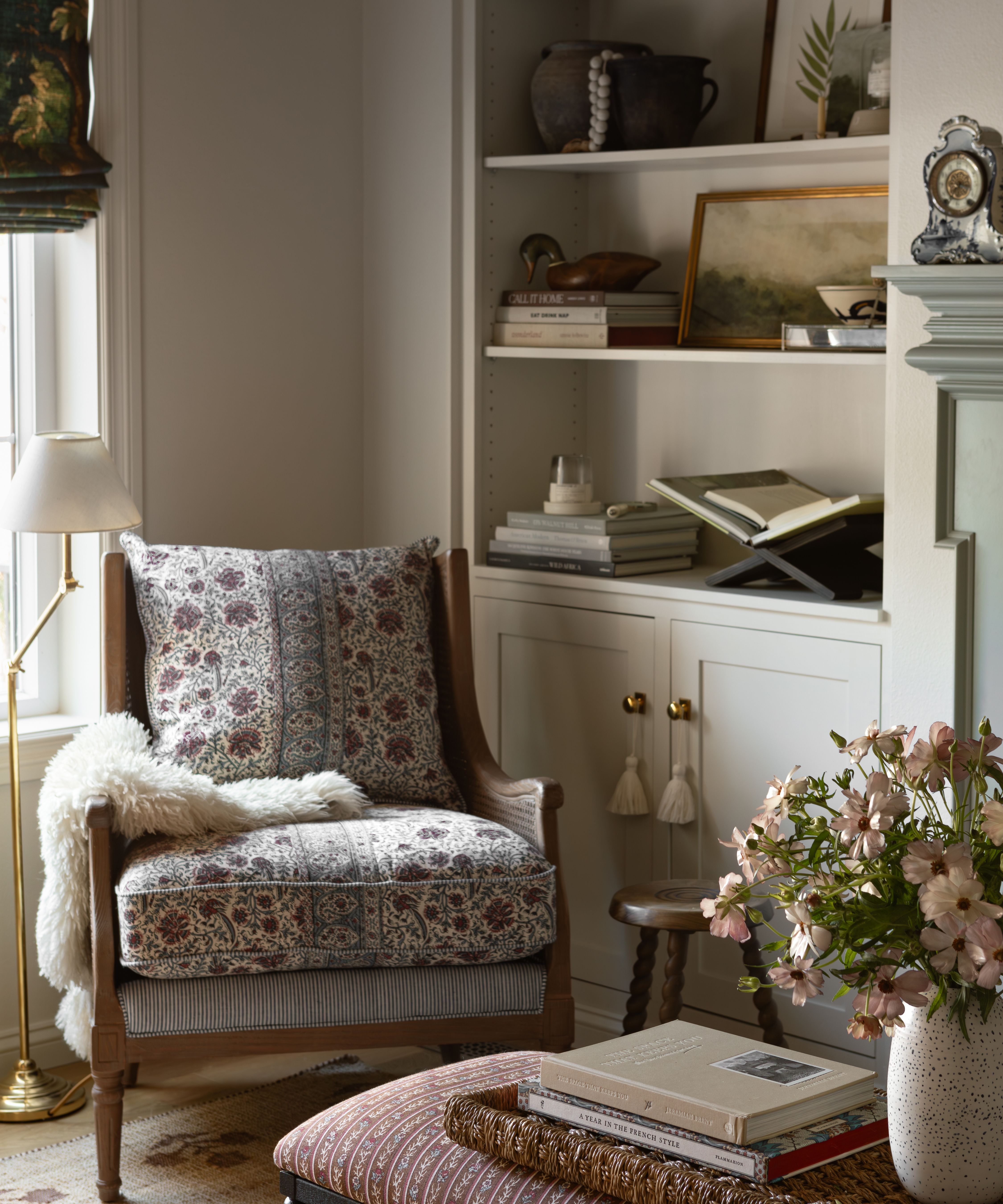 a corner of a neutral living room in a carriage house with built in bookcases, a window with a tapestry print blind and a floral upholstered wooden armchair styled with a floor lamp and bobbin stool