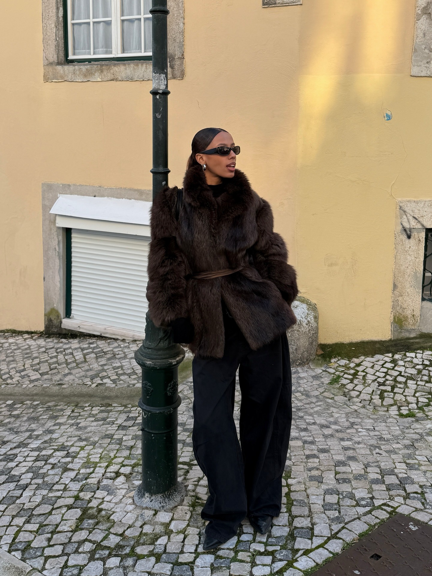 Fashion person Elise Sgn whaos what to wear in February. She wears a brown faux fur coat, black barrel legged jeans and pointed toe bots. She finishes here look with huggie hoops and oversized sunglasses. This image was taken from her Instagram recently.