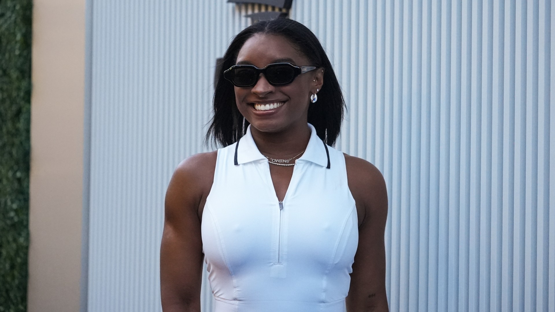 a black woman with dark hair wearing a sleeveless white shirt and black sunglasses smiling in front of a white background