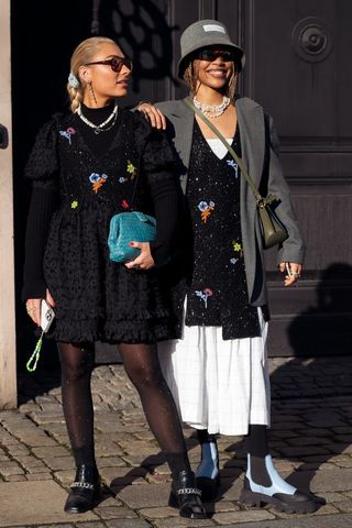 Maria Wos wearing black dress with embroidery and blue bag and Petra Henriette Rufi wearing long white dress, black vest with embroidery, grey coat, grey bucket hat and Ganni bag outside A. Roege Hove in Copenhagen fashion week Autumn/Winter 2022 on February 02, 2022 in Copenhagen, Denmark.