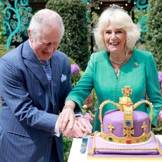 King Charles and Queen Camilla laugh while cutting a crown shaped cake