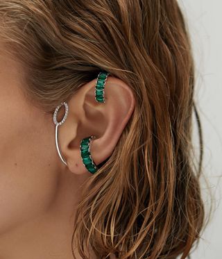 Woman's earring for unpierced ears by Nomis. Two ear rings with green gemstones clipped onto the side and top of the ear and a silver one with diamonds on curving around the front of the ear.