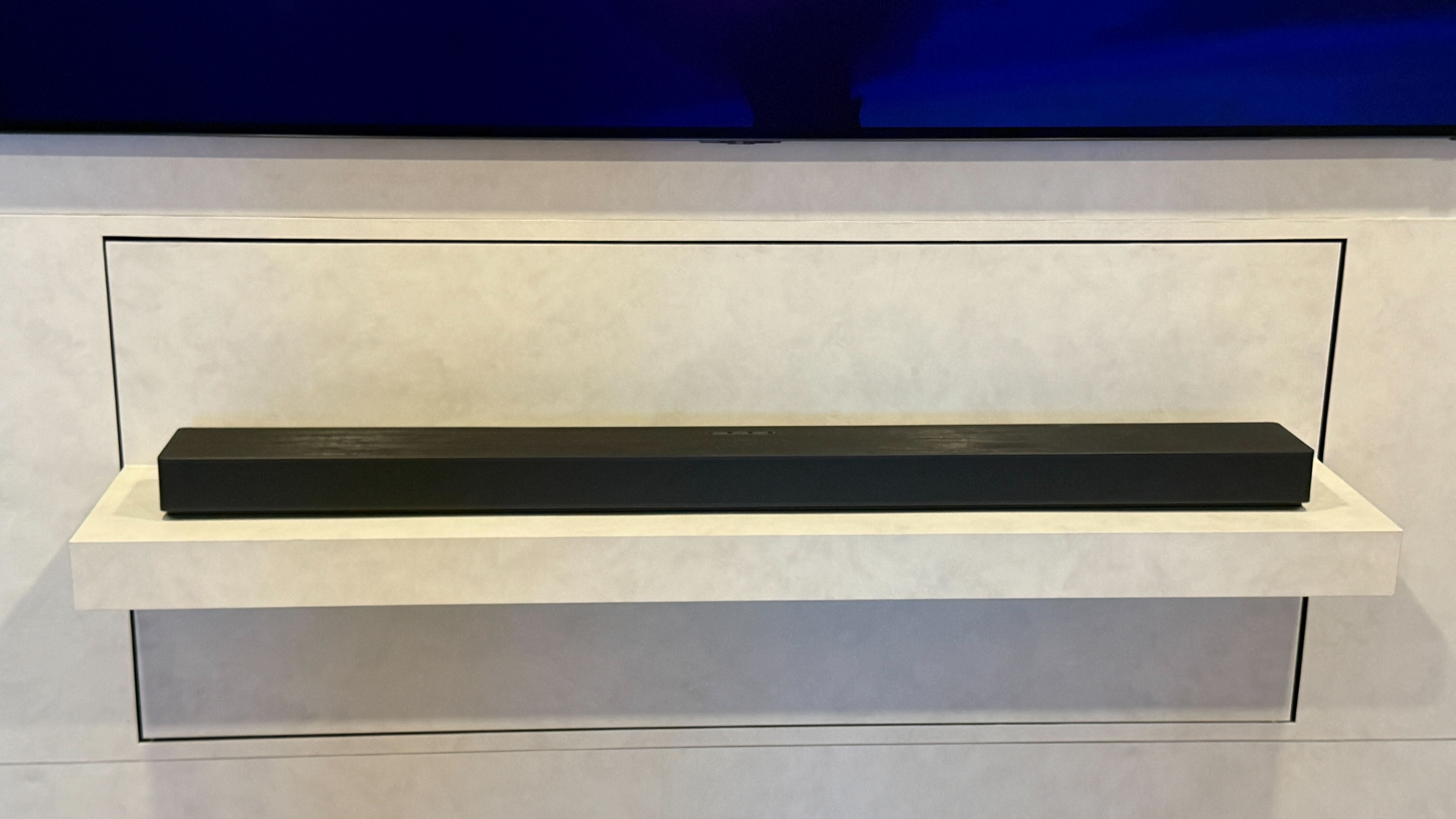 Samsung's HW-QS90H sitting below a wall-mounted TV on a white backdrop