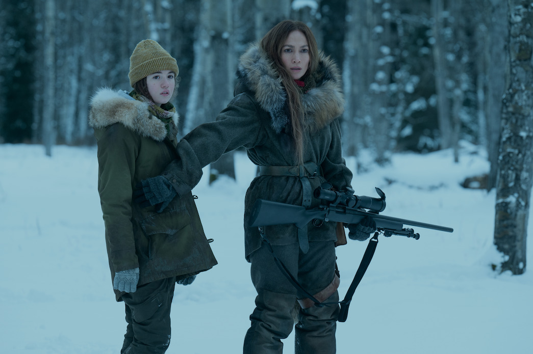 (L, R) Lucy Paez und Jennifer Lopez, die eine Waffe in der Hand hält, sind in den winterlichen Wäldern in „The Mother“.