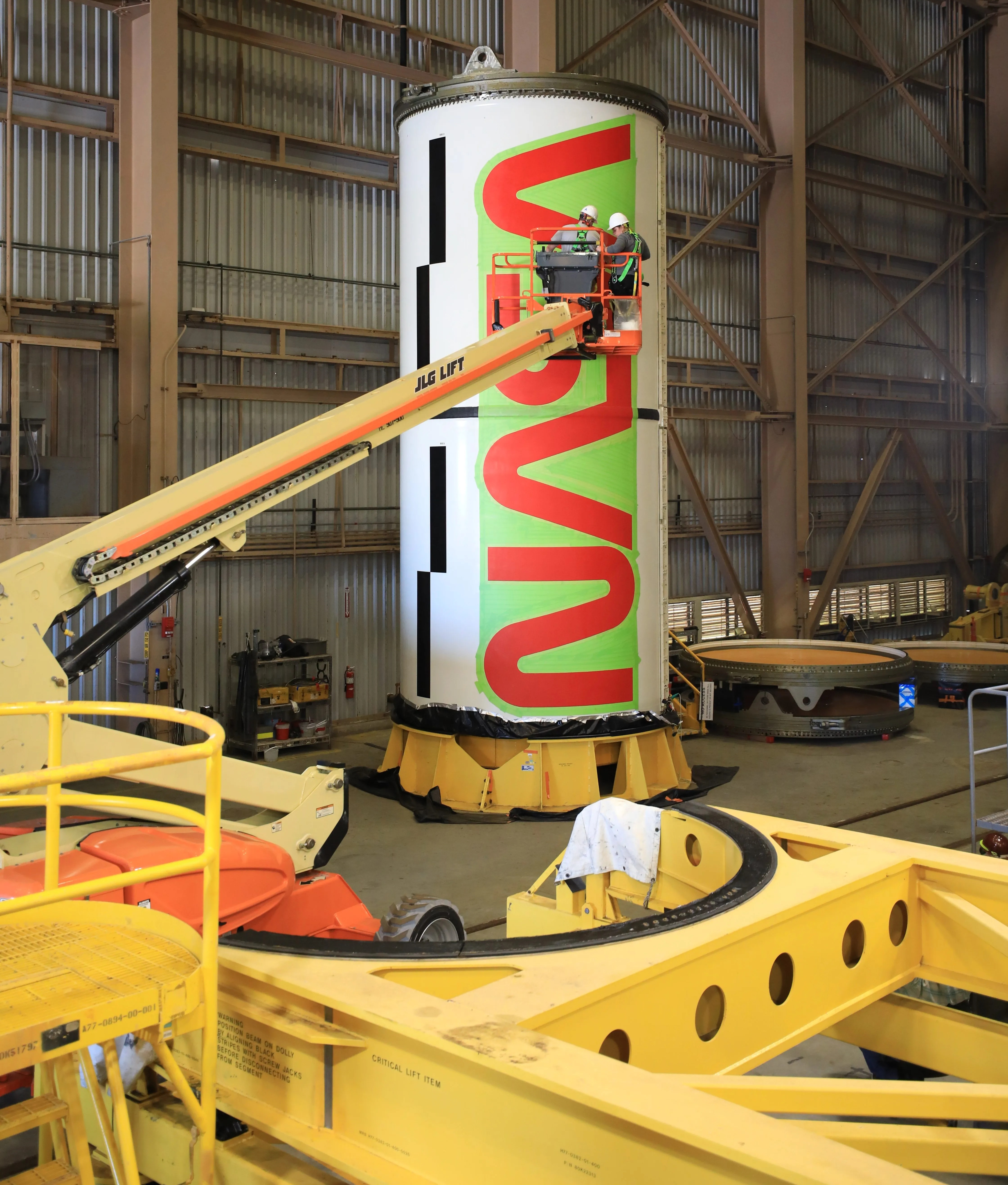 a faraway view of a rocket with a big nasa logo on it. in front are workers on a crane posting it on. in foreground is equipment