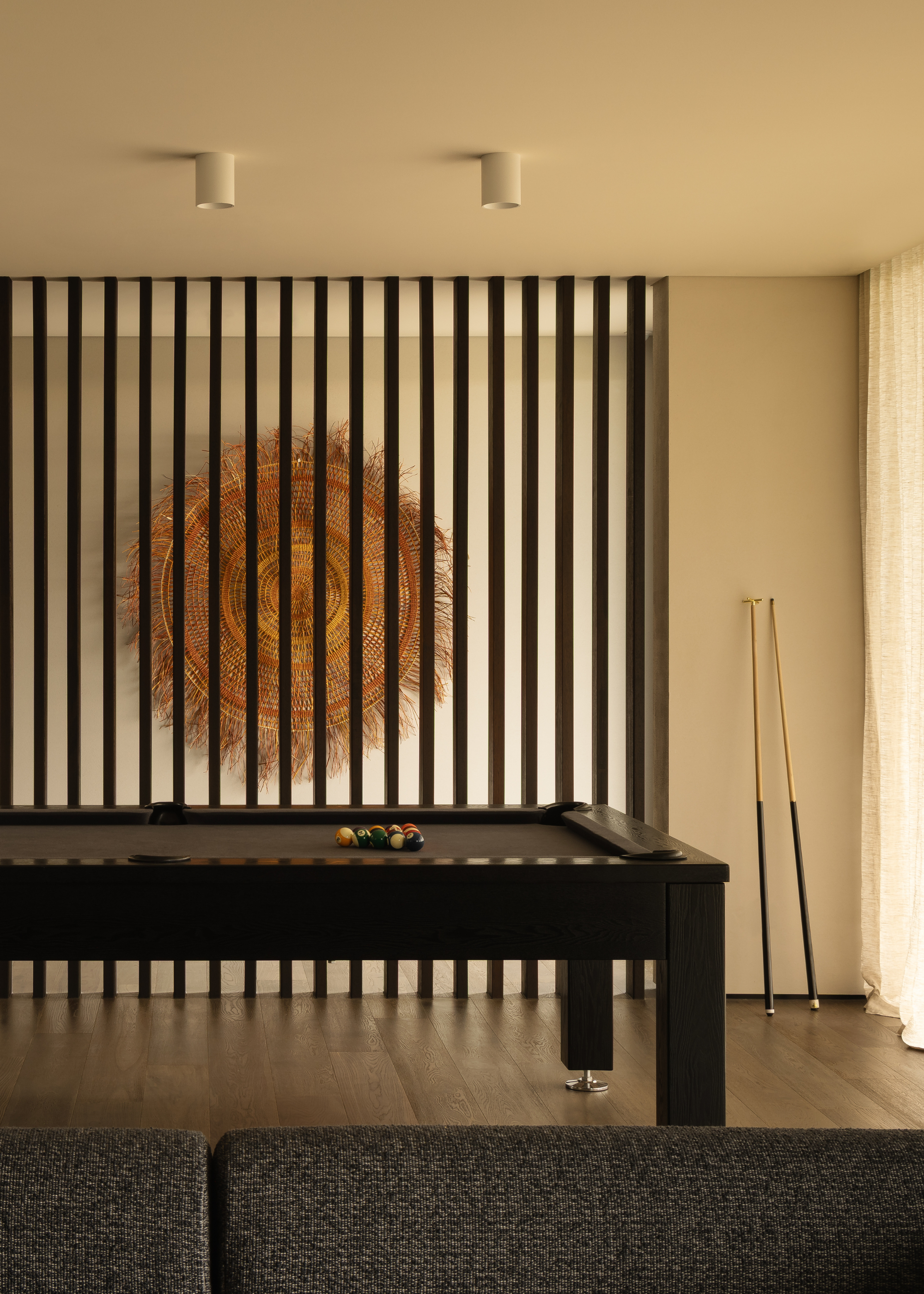 A games room with a black pool table, cues leaning on the wall, a slatted wood wall, and sheer window treatments