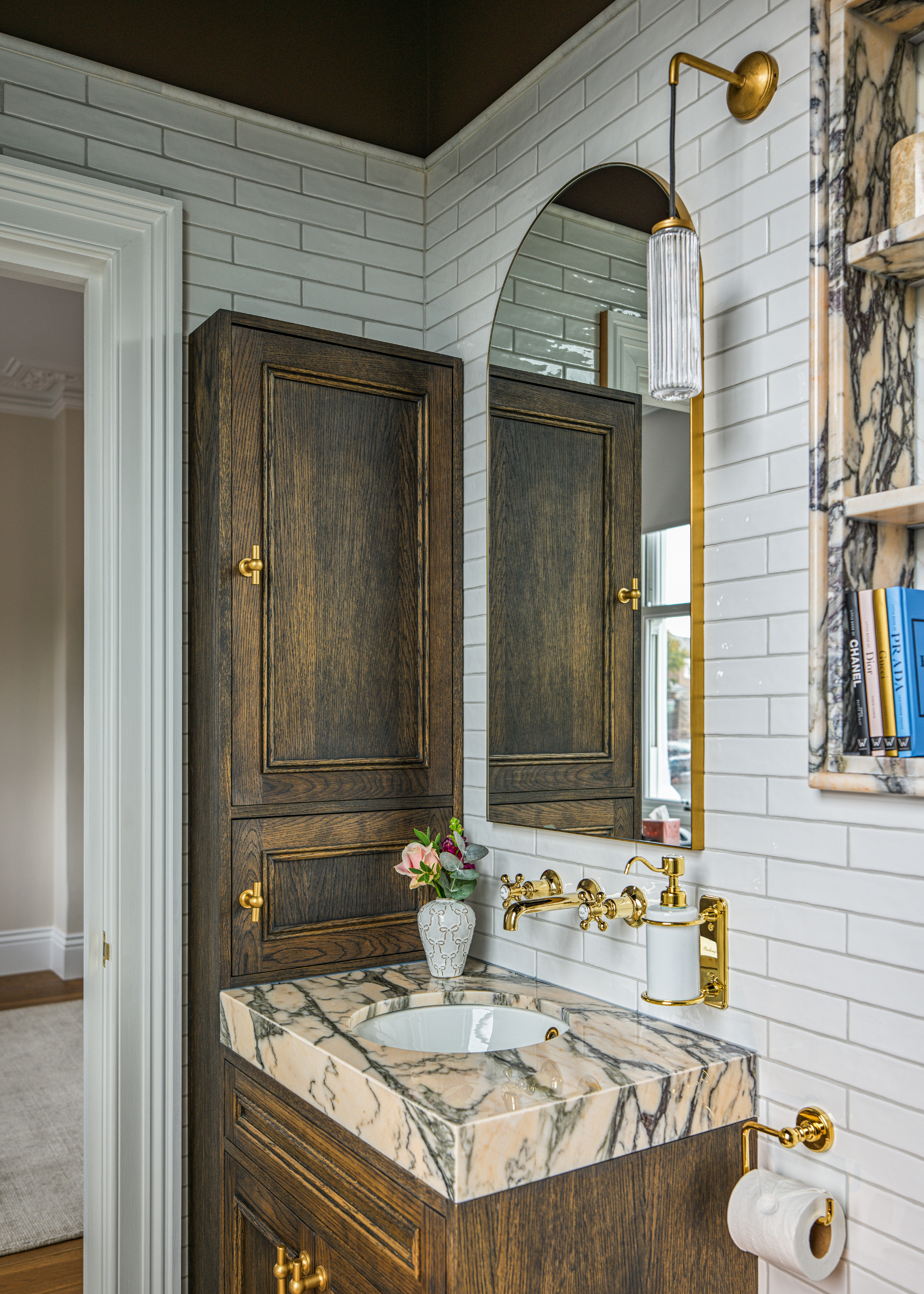 A modern take on a classic traditional bathroom featuring heavily veined pink and grey marble, white tiles, dark cabinetry and brushed brass fixtures and fittings.