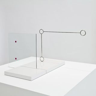 A piece of transparent perspex with two red dots, in front of an object constructed from two silver rods connecting three silver rings