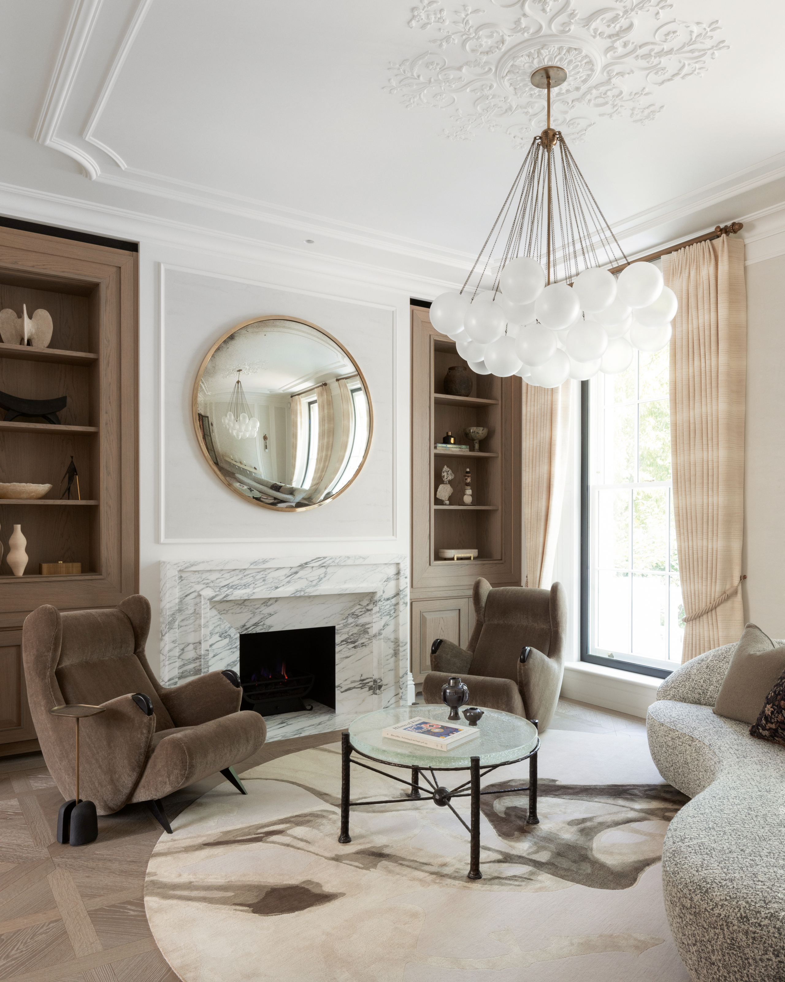 White living room with wooden built-in joinery either side of a white marble staircase, brown armchairs, grey curved sofa, glass and black-legged coffee table, smokey rug and wood parquet flooring
