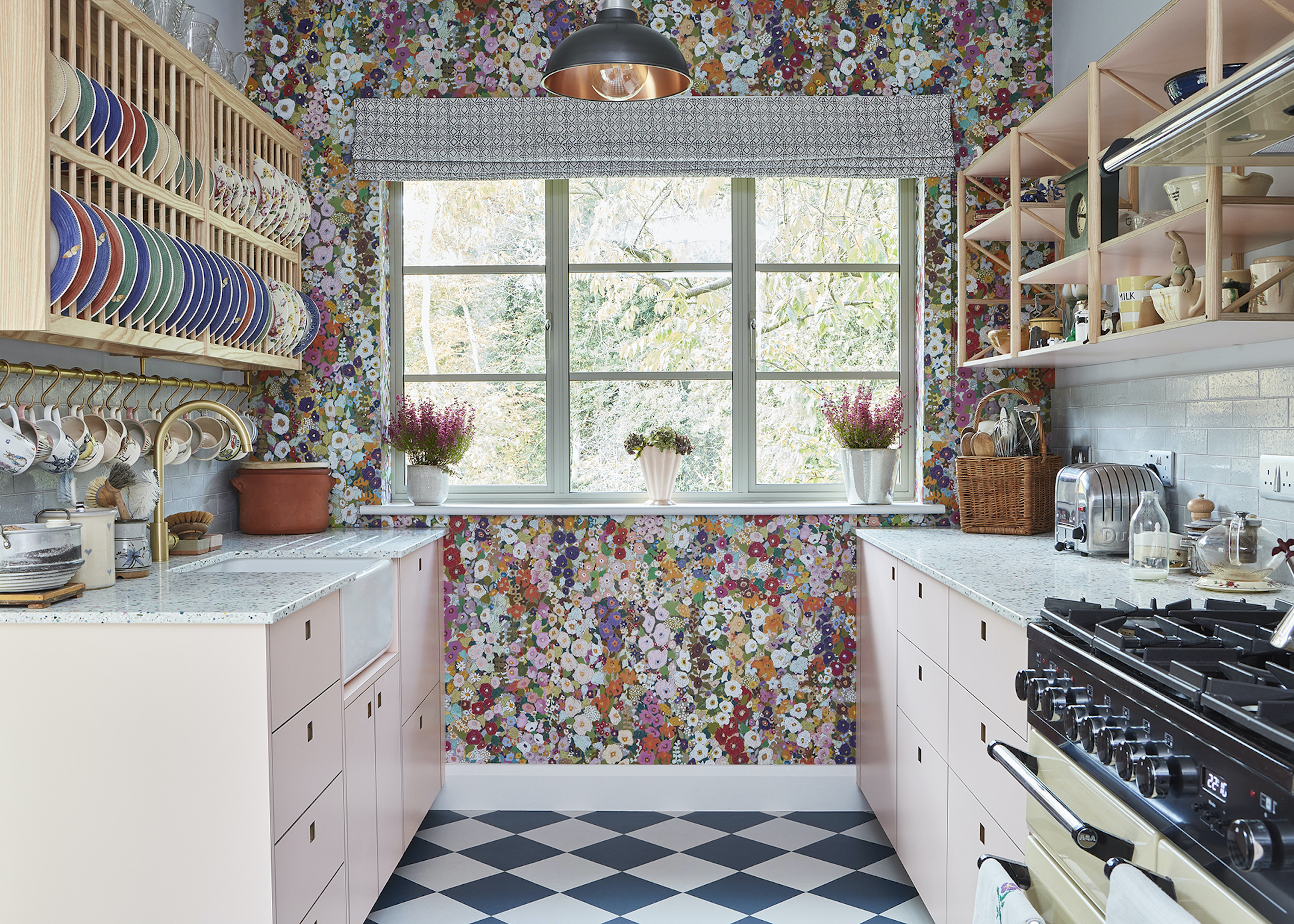 Pink kitchen with floral wallpaper, chequerboard flooring, white stone countertop, roman shade, and plate shelf and hanging mugs, with open shelving on the other side
