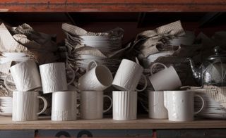 White ceramic cups with textured surfaces by Martino Gamper, with stacked plated seperated by papers behind in a cupboard