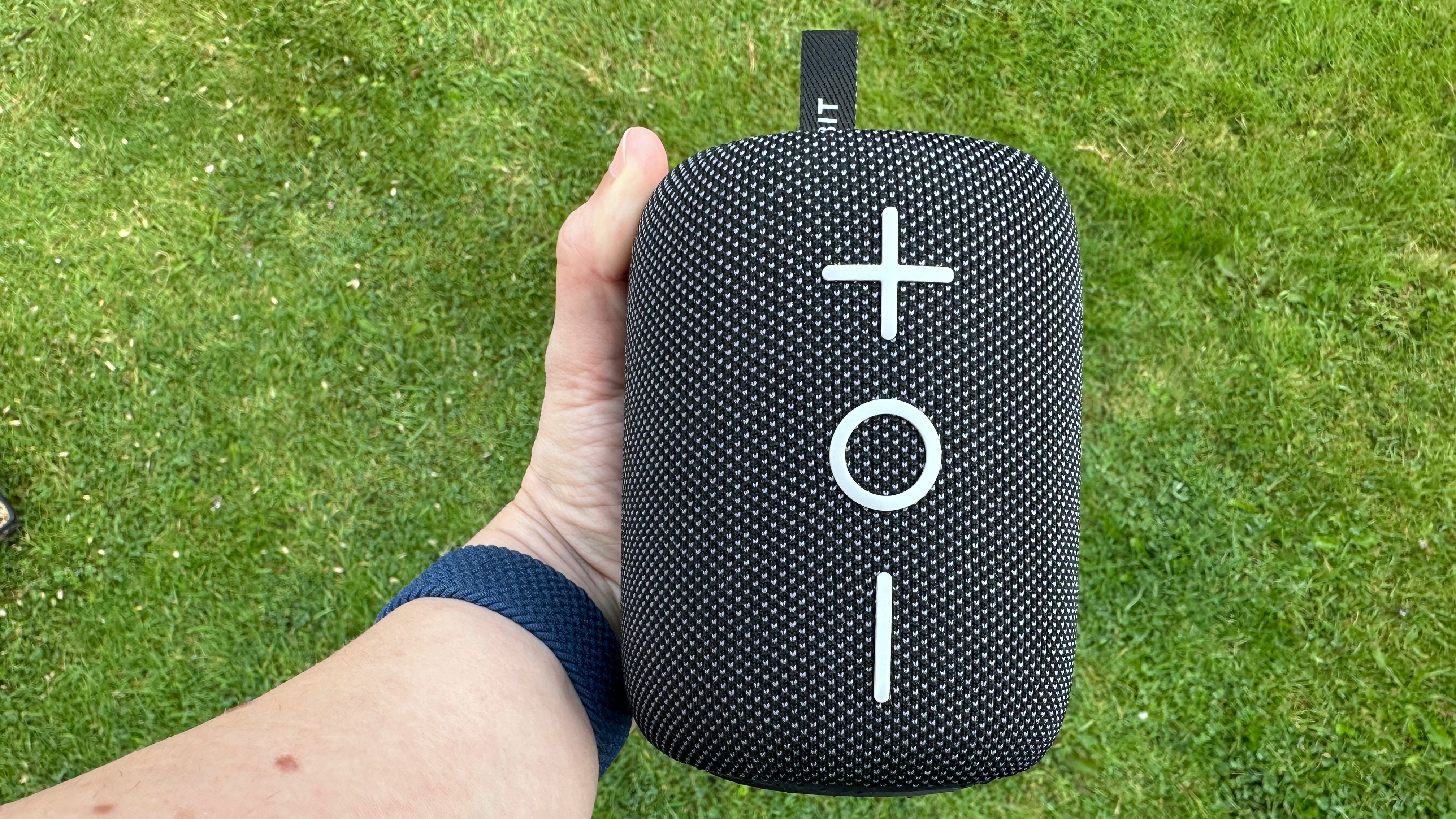 Tribit Stormbox Mini Plus Bluetooth speaker, held in a reviewer's left hand, above a grass surface